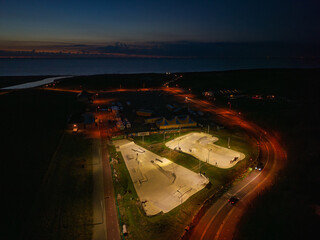 Aerial night view of skatepark brightly lit concrete ramps and bowls stand out against dark surroundings, nearby roads and vehicles, distant city lights shimmer across water under night sky.
