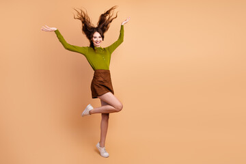 Joyful young woman jumps in the studio wearing a green top and brown skirt against a beige...