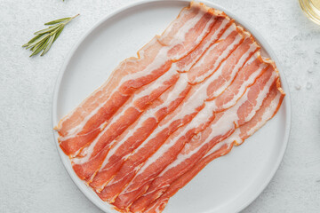 Uncooked bacon strips on white plate with rosemary and seasoning