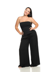 Fototapeta premium Studio shot of smiling woman with dark hair wearing black strapless top and wide pants posing with tilted head and hands on hips, full body, on white background.