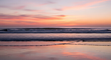 A serene ocean sunset with gentle waves reflecting the vibrant pink and orange hues of the sky on a sandy beach
