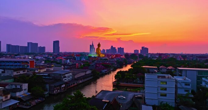 An aerial view of Bangkok at dusk reveals the iconic golden Buddha statue illuminated by twilight, surrounded by the city&rsquo;s modern skyline, temples, and the tranquil waters of Khlong Phasi Charoen.