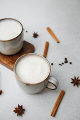 Two cups of warm frothy beverage with star anise and cinnamon sticks on a light textured surface.