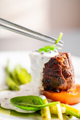 A gourmet dish of seared meat with creamy sauce is being meticulously garnished with a small green herb using tweezers, showcasing a focus on fine dining and culinary detail.