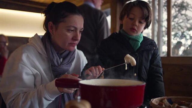 Mother and son sharing fondue meal in rustic restaurant, focused on dipping bread into hot melted cheese, warm family connection and cozy winter atmosphere