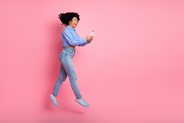 Energetic young woman joyfully leaping while holding a phone, showcasing a stylish casual outfit against a vibrant pink backdrop.