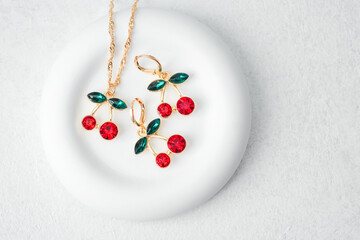 Elegant cherry earrings and necklace set with red gemstones and green leaves placed on white round plate