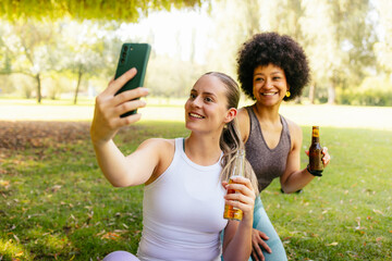 Two smiling friends relaxing in park after workout, taking selfie and drinking beer