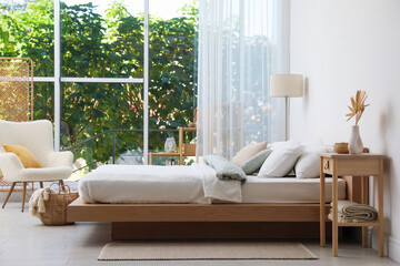 Cozy large bed with linens, furniture, houseplants and lamp near window in bedroom. Interior design