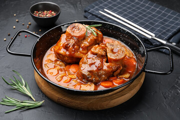 Tasty beef ossobuco with carrot, mushrooms and rosemary in frying pan on dark textured table, closeup