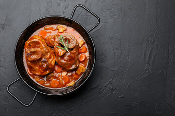 Tasty beef ossobuco with carrot, mushrooms and rosemary in frying pan on dark textured table, top view. Space for text