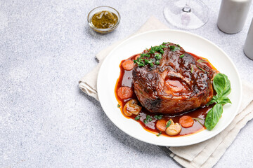 Tasty beef ossobuco with carrot, mushrooms and herbs on grey textured table, closeup. Space for text