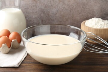 Liquid dough in bowl, whisk and ingredients on wooden table, closeup