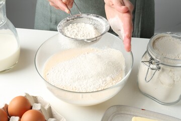 Making batter (liquid dough). Woman sieving flour into bowl at white table indoors, closeup