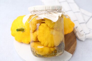 Pickled pattypan squashes in glass jar and fresh vegetables on white table, closeup