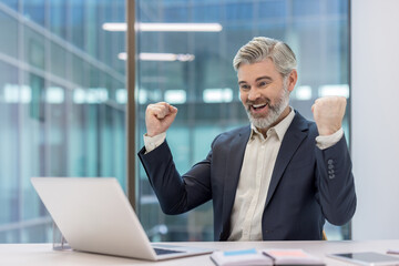 Mature businessman raising fists in triumph, celebrating financial achievement and online success...