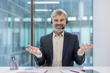 Smiling mature businessman with headset and microphone gesturing during a video call in a modern office, representing customer support, virtual consulting or telemarketing services