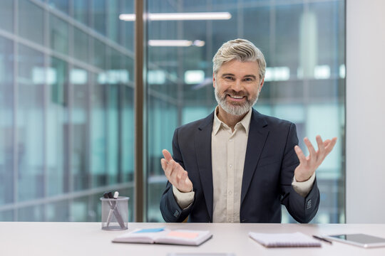 Mature executive, a confident businessman with a grey beard, smiling at the camera and gesturing with his hands during a virtual meeting or online presentation from a modern office desk