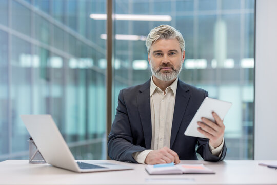 Confident mature businessman holding a digital tablet while sitting at a desk in a modern office, representing professionalism and using technology for business