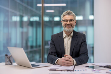 Experienced senior businessman wearing glasses and a suit, sitting confidently at a desk with a laptop and documents, smiling at the camera in a contemporary office setting