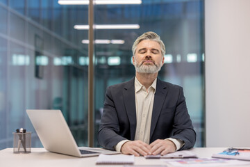 Mature businessman with eyes closed and hands clasped, practicing mindfulness and deep breathing at a modern office desk to relieve stress and restore calm during the workday