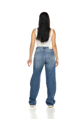 Fototapeta premium Studio shot of young woman with long dark hair wearing white tank top, blue jeans and sneakers standing rear view full length on white background.