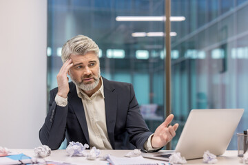 Mature businessman experiencing stress and frustration while working on laptop, surrounded by crumpled papers, struggling with a challenging task or creative block