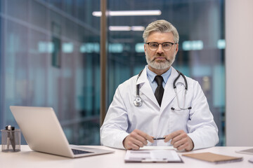 Fototapeta premium Experienced male physician in lab coat with stethoscope sits at desk with laptop and documents, looking at camera, projecting confidence and professionalism in a modern clinic office
