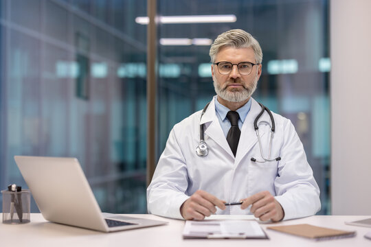 Experienced male physician in lab coat with stethoscope sits at desk with laptop and documents, looking at camera, projecting confidence and professionalism in a modern clinic office - Powered by Adobe