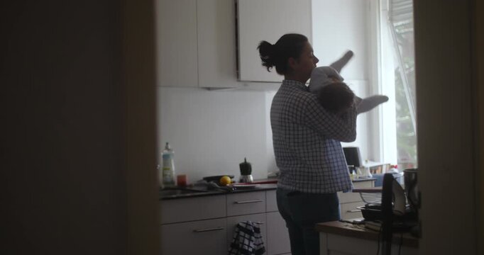 Mother holding baby in kitchen during quiet morning light, tender everyday moment showing care, warmth, and the soft rhythm of domestic life