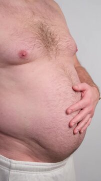 Overweight man's torso in profile view slapping his large belly against a white background, highlighting problems with excess weight, poor diet, and an unhealthy sedentary lifestyle