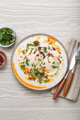 Turkish eggs cilbir, a delicious and colorful breakfast with poached eggs on yogurt, seasoned with spices and fresh herbs, arranged on a light wooden table