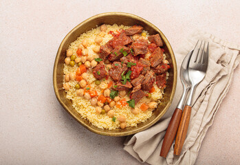 Traditional dish of Maghreb cuisine, a Moroccan couscous with lamb or beef, vegetables, chickpeas, and peas in a rustic bowl with cutlery on beige background