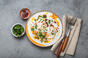 Turkish eggs cilbir, a delicious and colorful breakfast with poached eggs on yogurt, seasoned with spices and fresh herbs, arranged on grey concrete background