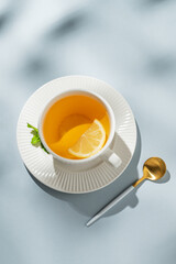 Hot tea with lemon, honey, and mint in a cup on a blue background with spoon and morning shadows.