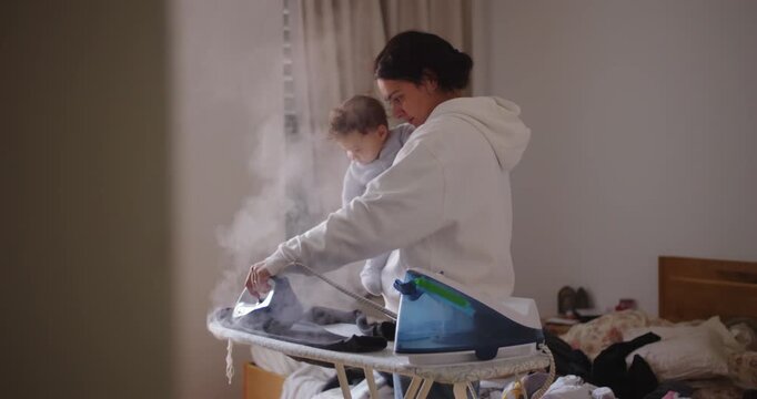 Mother holding baby while ironing clothes in messy bedroom, capturing the tender chaos of daily family life and the quiet strength of motherhood in routine care