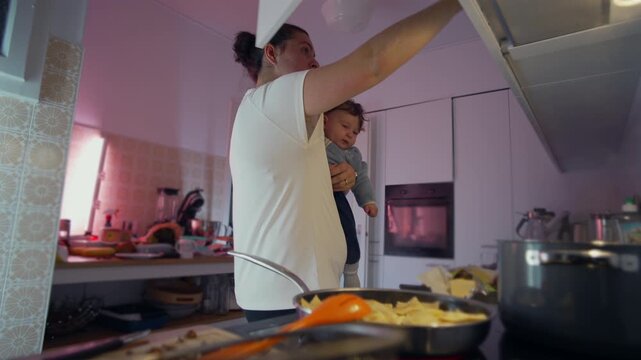 Mother holding baby while putting items away in kitchen cabinet, everyday family moment showing balance between nurturing, organization, and domestic life at home