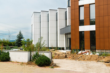 Contemporary office building under construction with contrast of white panels and brown wood-look facade panels, showing rough landscaping and dirt piles