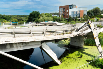 A concrete road bridge, destroyed as a result of hostilities, collapses over a ditch of stagnant green water, symbolizing the devastating impact of war