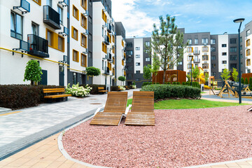 Wooden sun loungers and a gravel relaxation area in the landscaped courtyard of a newly built, colorful modern apartment block complex under a sunny sky