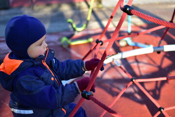 Close-up of little boy in blue snowsuit holding red playground ropes, exploring climbing structure...
