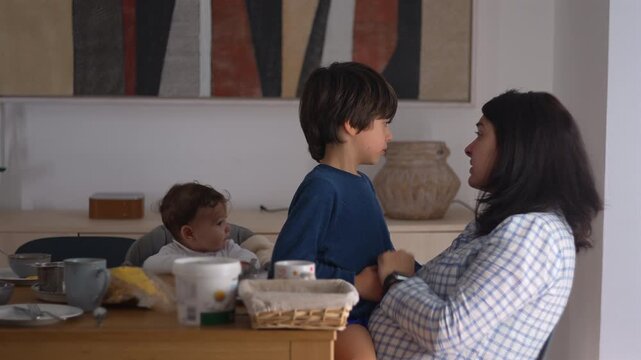 Mother talking to son while holding him on her lap as baby looks on, guiding and educating him about misbehavior with calm authority and emotional connection at home