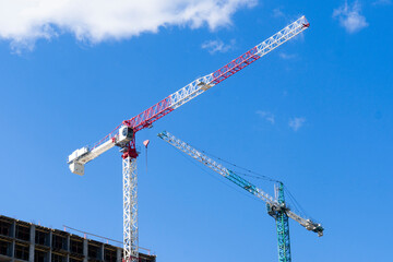 A pair of towering red and green construction cranes with long jibs dominate the frame, working on a new building under a clear blue sky with white clouds