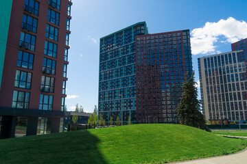 Contemporary urban residential complex featuring colorful high-rise apartment buildings set against a clear blue sky, with a bright green manicured lawn in the foreground
