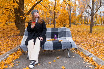 Young woman in a coat and beret sitting on a bench covered with a plaid blanket, enjoying a coffee in a beautiful autumn park