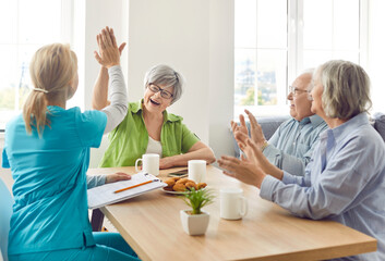 Obraz premium A nurse in a nursing home cheerfully communicates with the elderly, close-up. Pensioners gesture is all right. An employee of a boarding house drinks tea with elderly guests. Retirement age