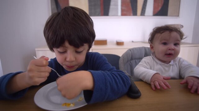 Older brother eating cereal while baby watches curiously from high chair, showing sibling interaction and early social observation during breakfast at home