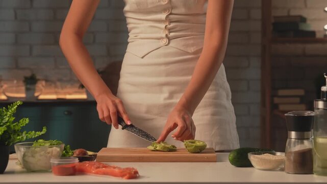 A woman prepares healthy food in her kitchen and slices an avocado. On the table are cucumber, fresh lettuce leaves, salmon, and spices. Healthy eating, eco friendly products.