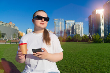 Young woman in sunglasses holding a phone and coffee cup while relaxing on green grass with modern skyscrapers in the background
