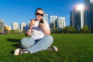 Young woman in sunglasses holding a phone and coffee cup while relaxing on green grass with modern skyscrapers in the background